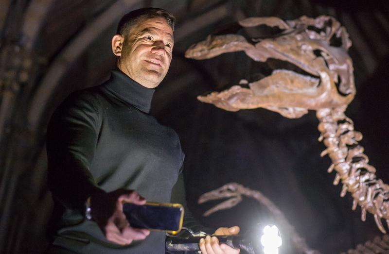 Steve backshall at OUMNH Man holding a flashlight and mobile phone in a dark space below the skeleton of a dinosaur